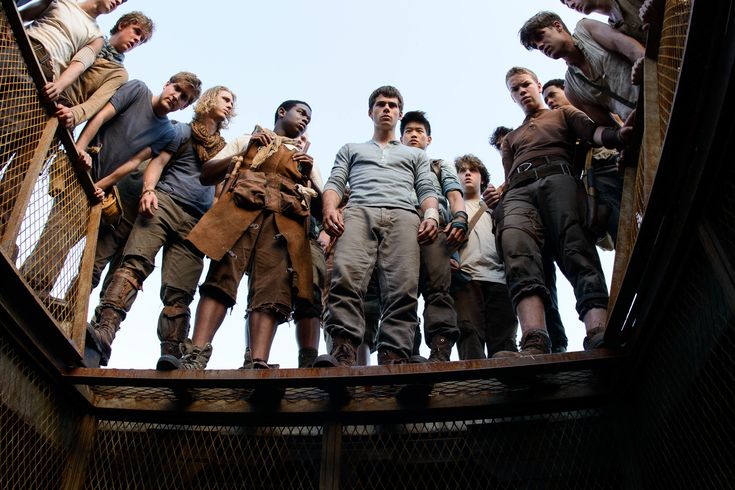 Cena do filme Maze Runner: grupo de jovens olhando para baixo através de uma grade metálica, em um ambiente cercado por estruturas enferrujadas, sob céu aberto.